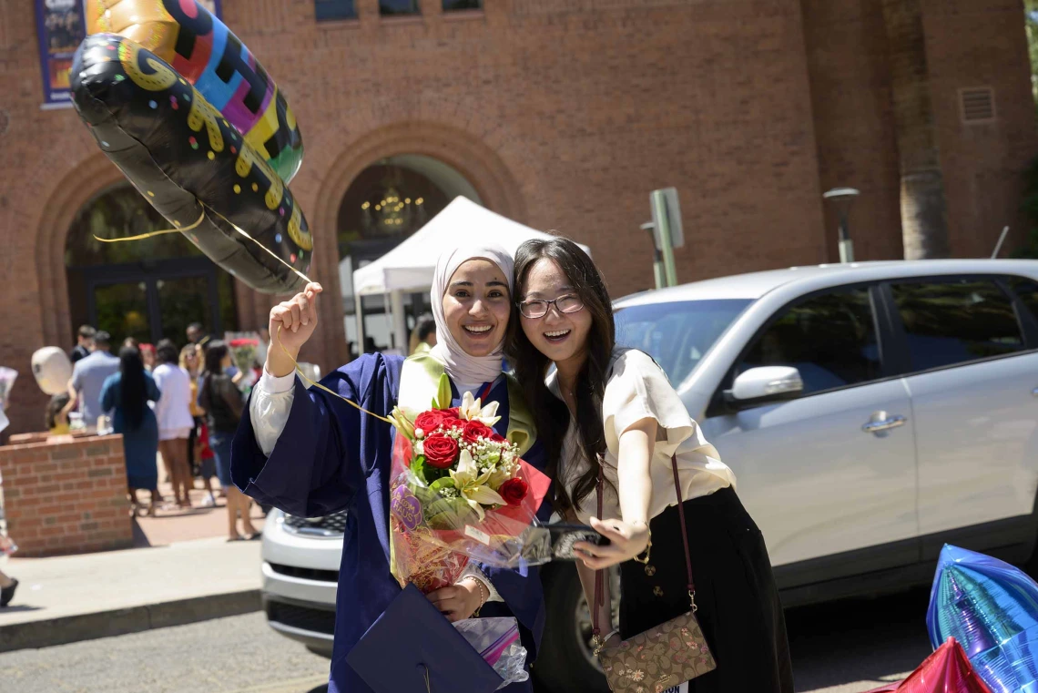 Two University of Arizona R. Ken Coit College of Pharmacy students, one wearing a graduation gown and holding a balloon and flowers, hug after the college’s graduation ceremony.