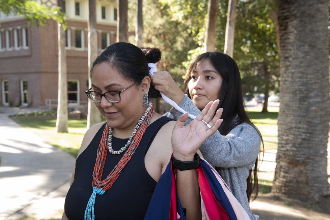 A University of Arizona Mel and Enid Zuckerman College of Public Health student wearing a large beaded necklace stands outside as her niece ties her hair in a bun.