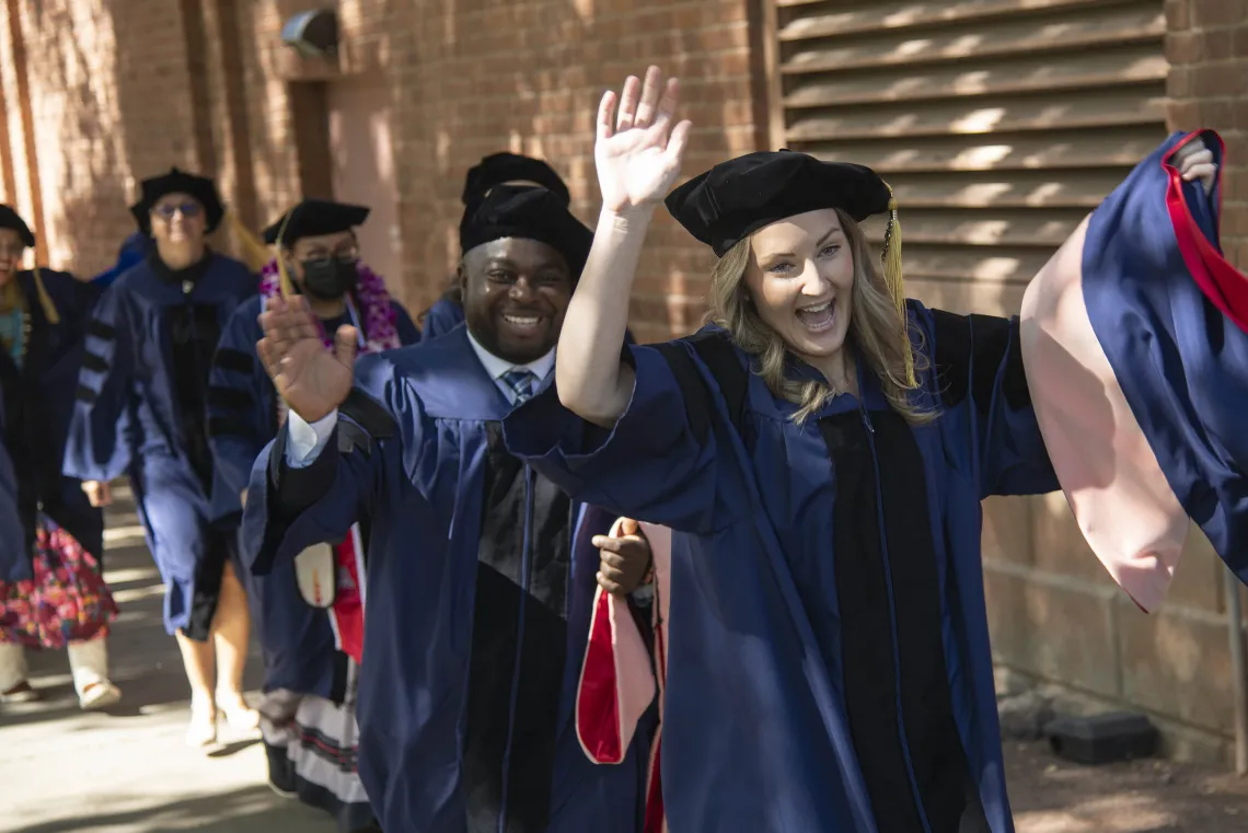 Several University of Arizona Mel and Enid Zuckerman College of Public Health students wearing graduation caps and gowns smile and wave as they walk outside. 