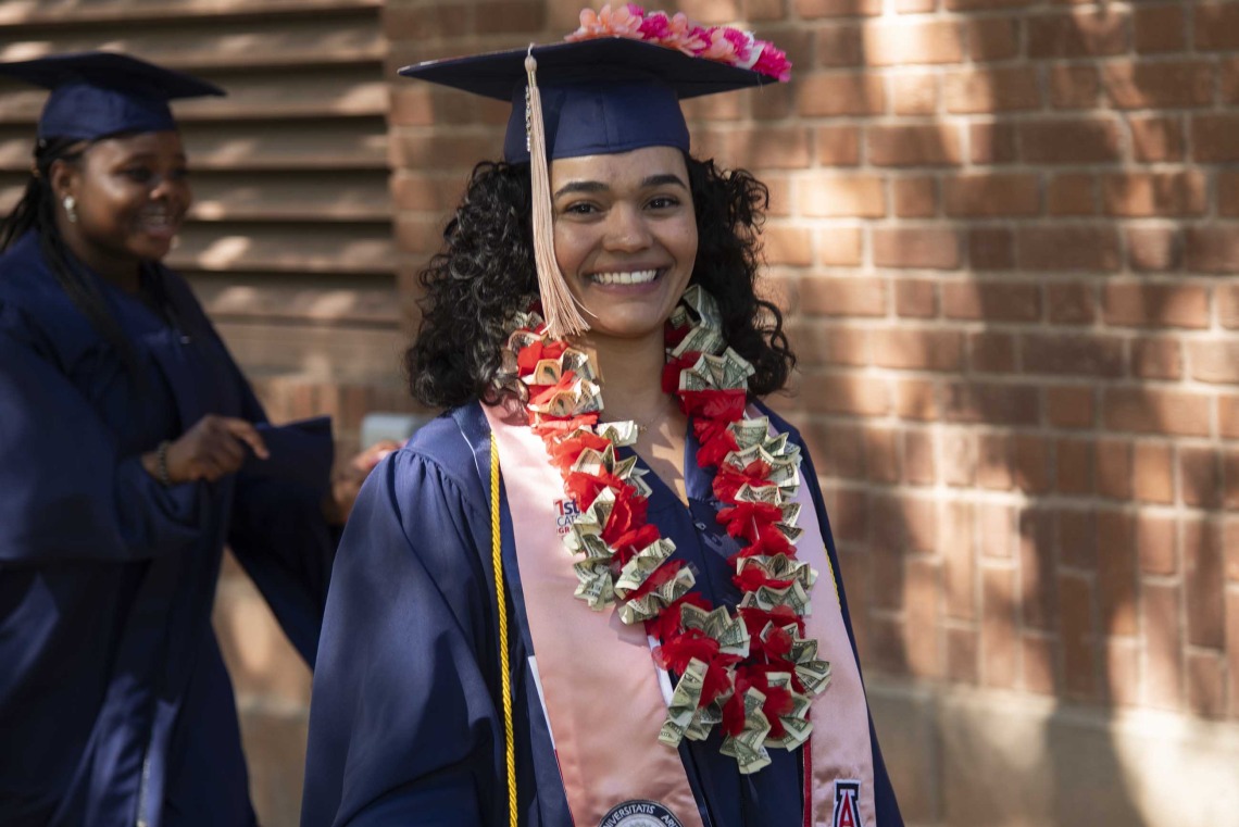 A University of Arizona Mel and Enid Zuckerman College of Public Health student wearing a graduation cap and gown smiles while walking outside. 