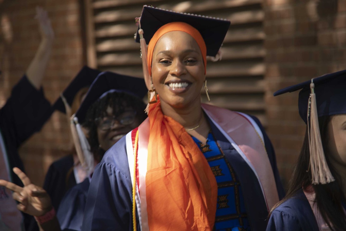 A smiling University of Arizona Mel and Enid Zuckerman College of Public Health student wearing a graduation cap and gown and an orange headscarf walks outside with other students.