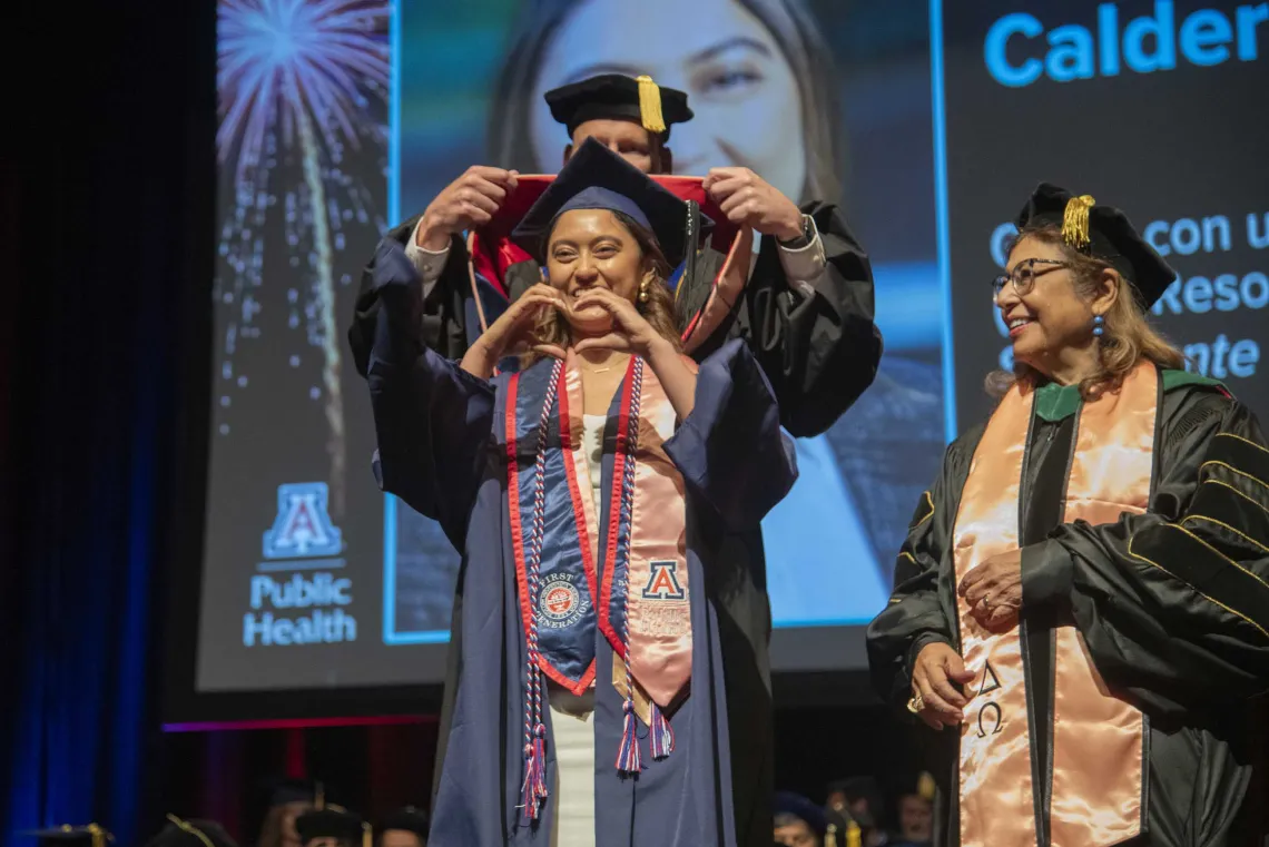 A University of Arizona Mel and Enid Zuckerman College of Public Health student makes a heart sign with her hands as she is hooded by a faculty member as another watches. All are wearing graduation regalia.