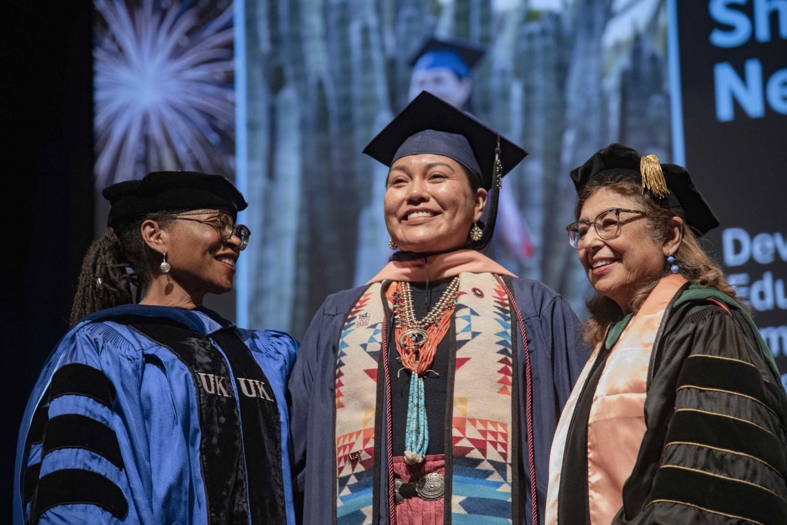 A University of Arizona Mel and Enid Zuckerman College of Public Health student smiles as she stands between two faculty members. All are wearing graduation regalia.