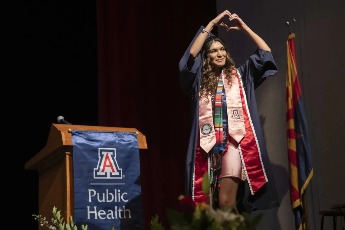 A University of Arizona Mel and Enid Zuckerman College of Public Health student makes a heart sign with her hands as she walks acroos a stage while wearing a graduation cap and gown.