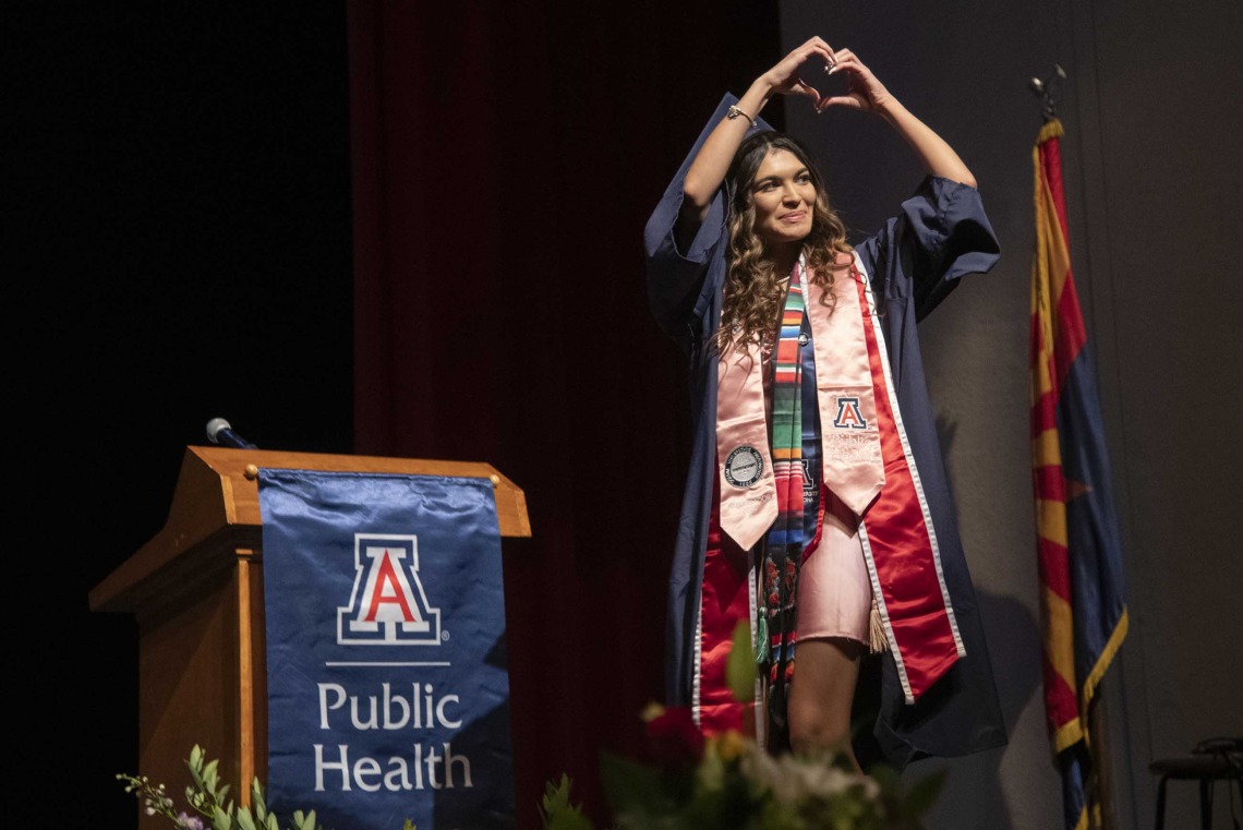 A University of Arizona Mel and Enid Zuckerman College of Public Health student makes a heart sign with her hands as she walks acroos a stage while wearing a graduation cap and gown.