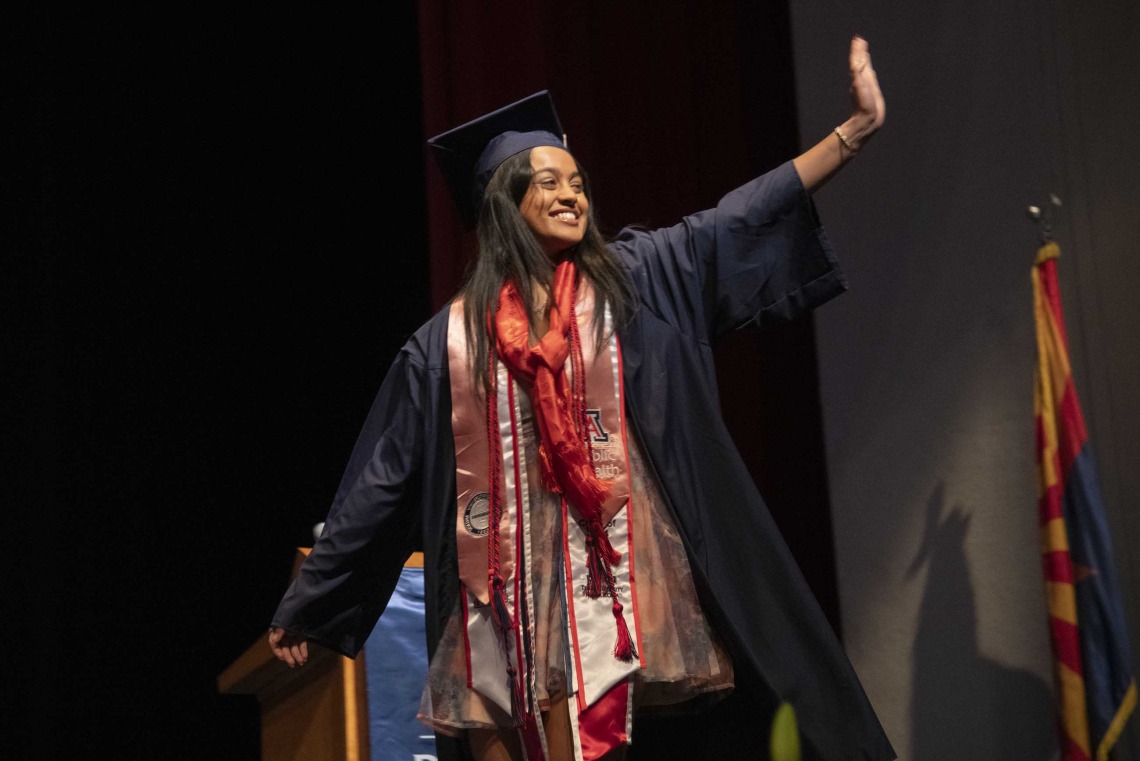 A University of Arizona Mel and Enid Zuckerman College of Public Health student, smiling and dressed in graduation regalia, waves while walking across a stage. 