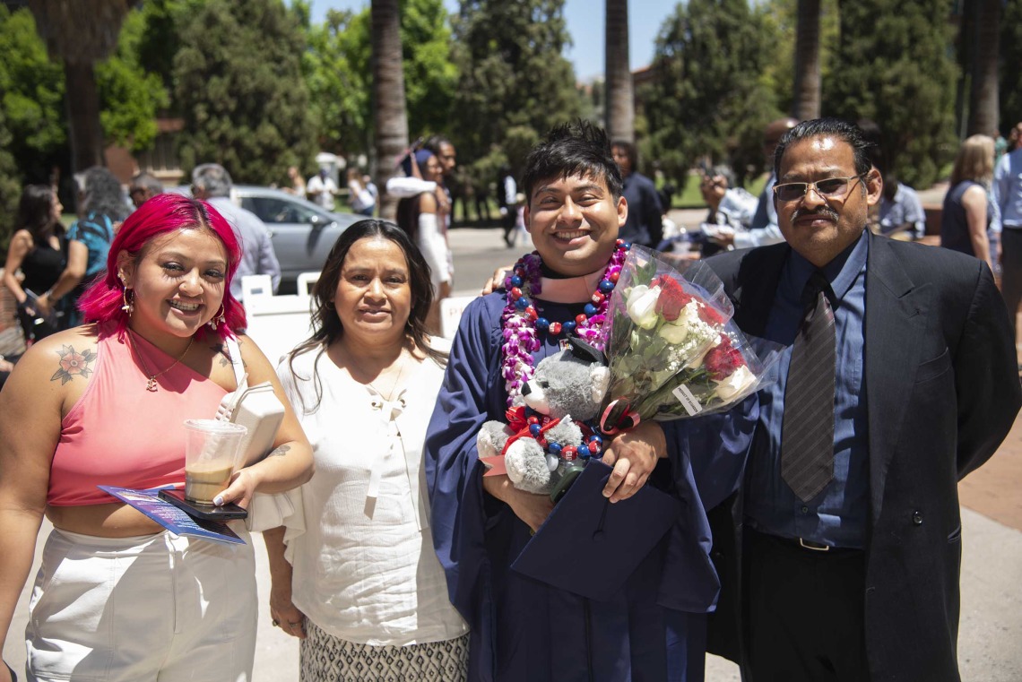 A University of Arizona Mel and Enid Zuckerman College of Public Health student smiles while holding flowers, flanked by three family members. The student is wearing a graduation gown. 
