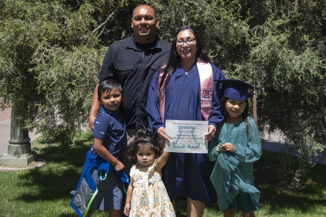A University of Arizona Mel and Enid Zuckerman College of Public Health student wearing a graduation gown holds up her diploma while standing with her family outside. 