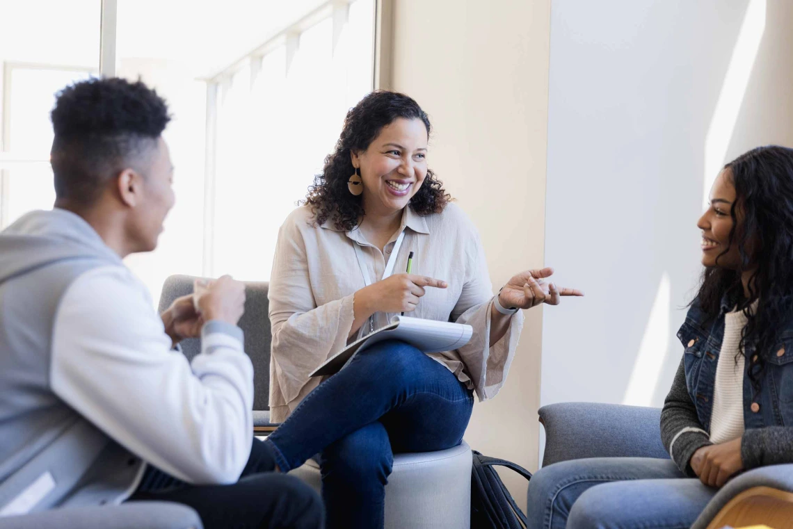 A woman with a notepad on her lap and a pen in her hand sits between two smiling college students. 