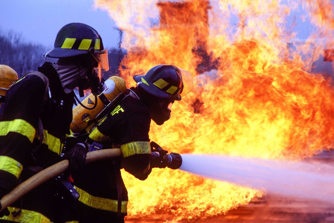 Two firefighters wearing protective gear and oxygen tanks spray water onto a blaze