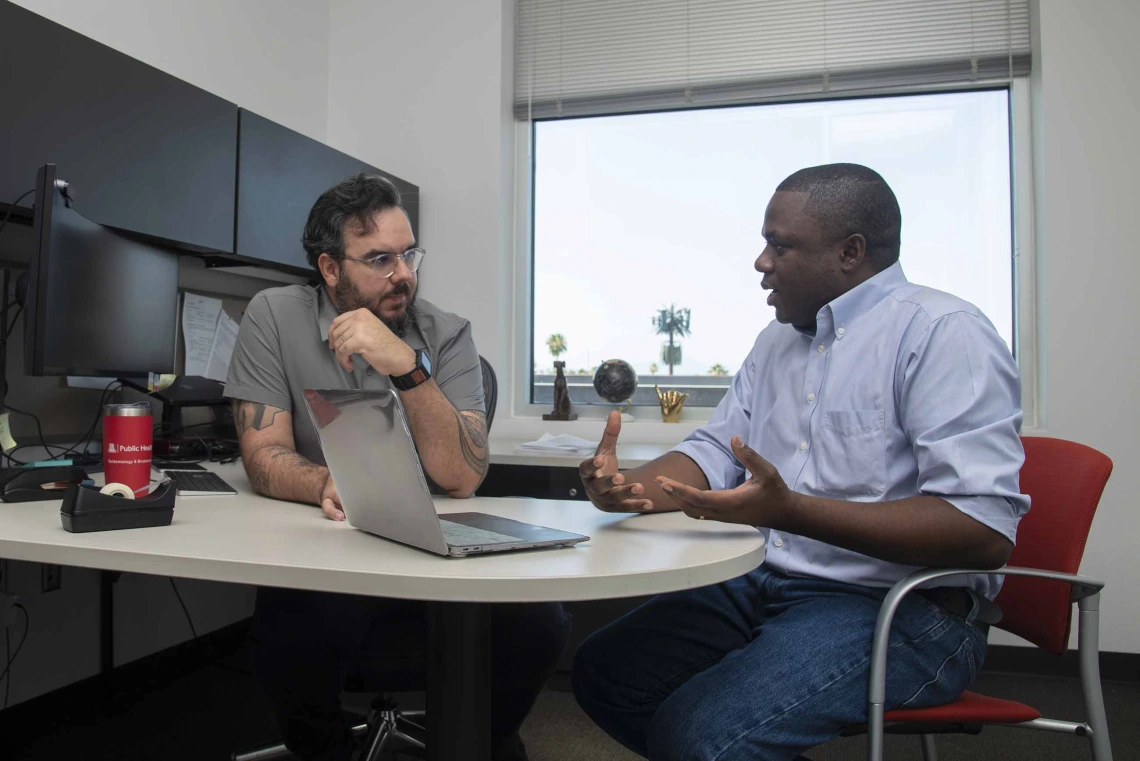 Two men sit in an office talking with an open laptop in front of them.