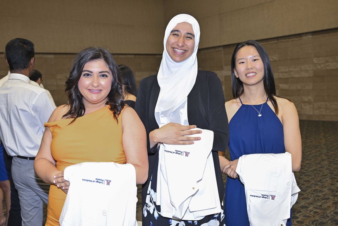 Three new University of Arizona College of Medicine – Phoenix students, with medical white coats draped over their arms, smile as they stand side by side.