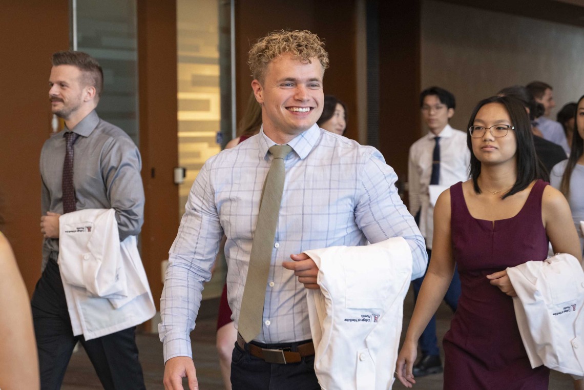 A new University of Arizona College of Medicine – Phoenix student dressed in a shirt and tie smiles as he holds a medical white coat over his arm.