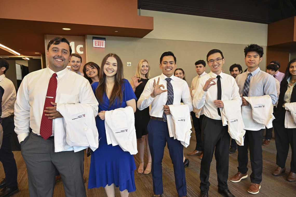A group of new U of A College of Medicine – Phoenix medical students dressed in shirts, ties and dresses, all with medical white coats draped over their arms, stand, smile and show the hand sign for the Arizona Wildcats. 