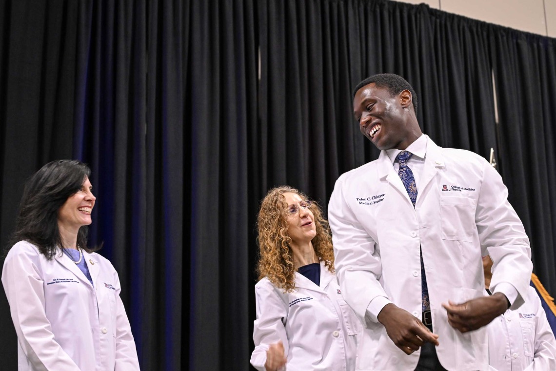 A new, male student at the University of Arizona College of Medicine – Phoenix smiles at two female professors on a stage. All are wearing medical white coats. 