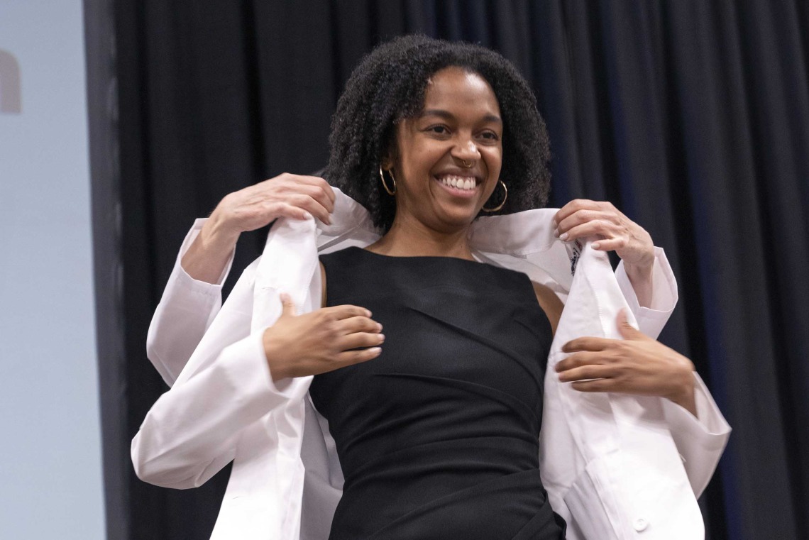 A smiling University of Arizona College of Medicine – Phoenix student in a black dress adjusts a medical white coat that is being put on her by a professor.