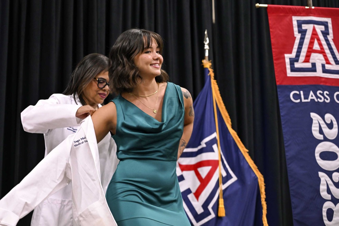 A smiling University of Arizona College of Medicine – Phoenix student in a green dress stretches out her arms as a professor puts a medical white coat on her.