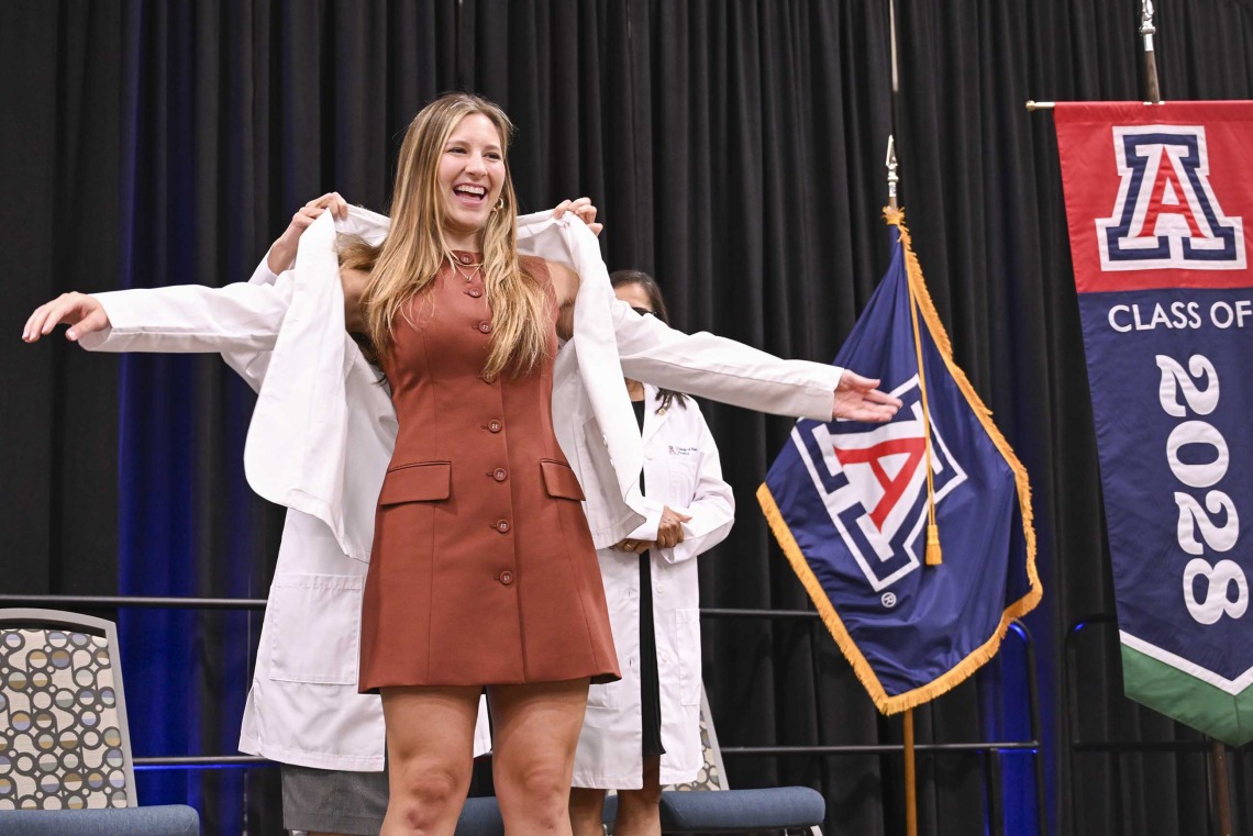 A new University of Arizona College of Medicine – Phoenix student in a brown dress stretches out her arms as a professor puts a medical white coat on her.