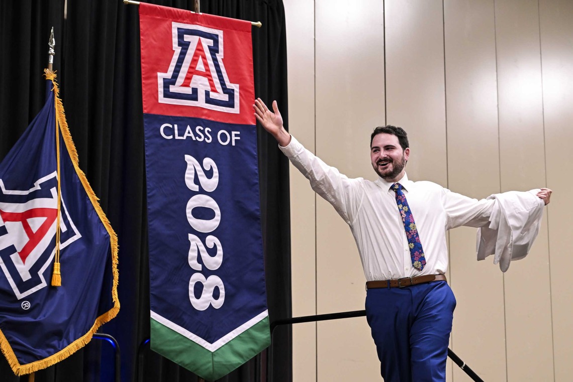A new University of Arizona College of Medicine – Phoenix student with a medical white coat draped over one arm extends the other arm outward as he smiles and walks onto a stage. Behind him is a  U of A banner that reads “Class of 2028.” 