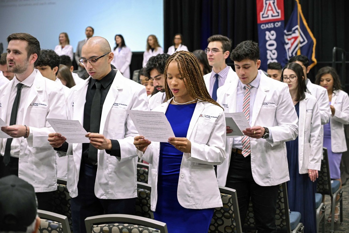 A large group of University of Arizona College of Medicine – Phoenix students stand in their white coats reading from printouts they are holding. 
