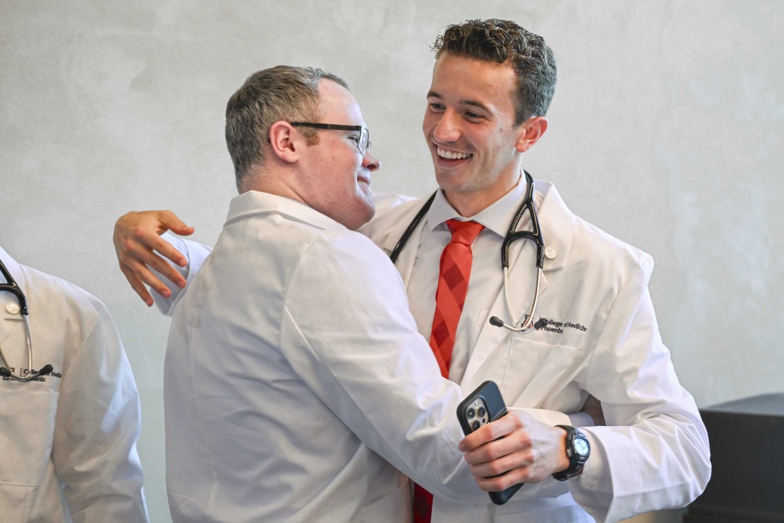 Two University of Arizona College of Medicine – Phoenix students wearing medical white coats smile as they hug. 