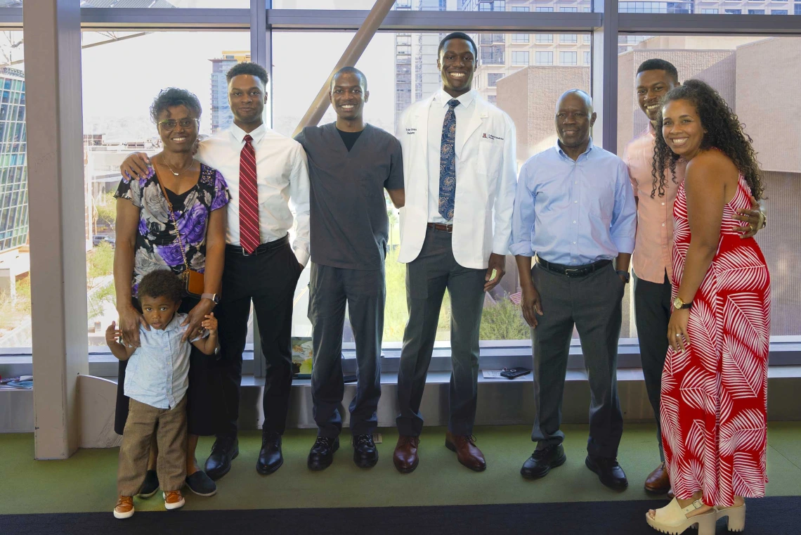 Several family members stand on each side of a University of Arizona College of Medicine – Phoenix student who is wearing a medical white coat. All are smiling.