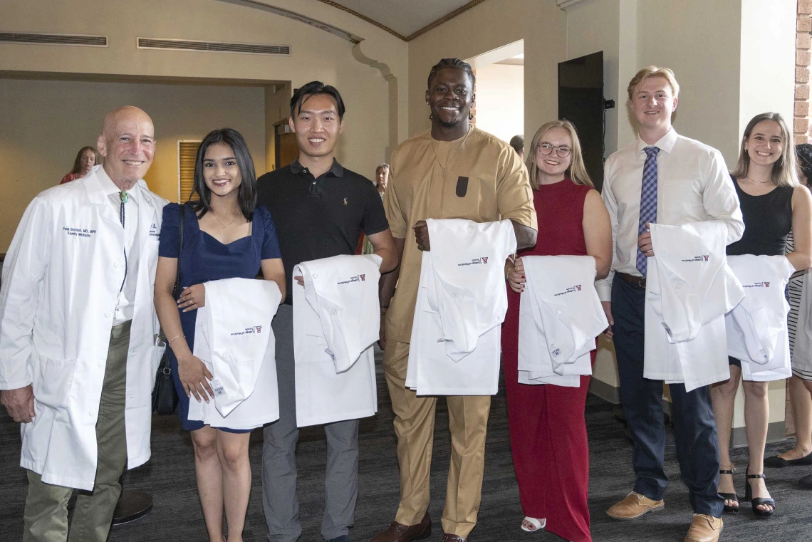 A University of Arizona College of Medicine – Tucson professor wearing a white medical coat stands with six new medical students, all with white coats draped over their arms. 