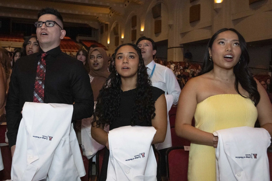 Three new University of Arizona College of Medicine – Tucson students with white coats draped over their arms stand in front of a large group of fellow students and recite their class oath.