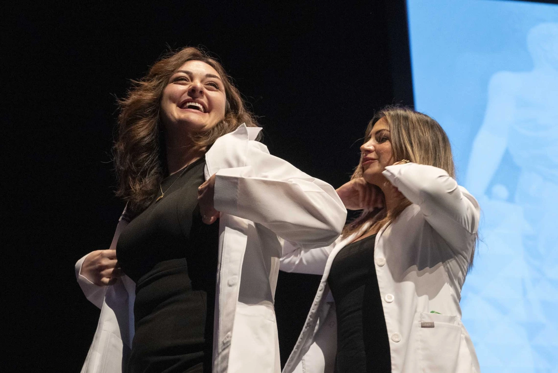 A smiling University of Arizona College of Medicine – Tucson student adjusts a medical white coat that was just put on her by a professor.