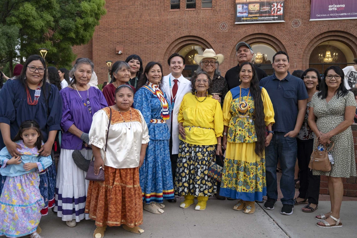 An incoming University of Arizona College of Medicine – Tucson student wearing a medical white coat stands with more than a dozen smiling family members outside near a red brick building. Several of the women in the group are wearing traditional Native American outfits.  