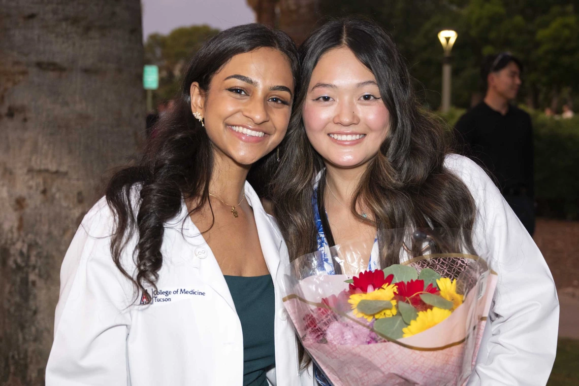 Two incoming University of Arizona College of Medicine – Tucson students wearing medical white coats stand together, smiling. One is holding flowers. 