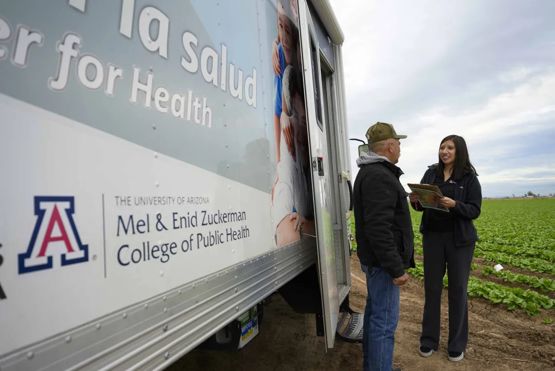 A University of Arizona Mel and Enid Zuckerman College of Public Health professor interviews a farmworker as they stand in a field next to a mobile health trailer. 