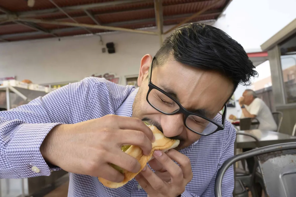 A medical student in a blue shirt bites into a Sonoran hot dog at an outdoor food stand. 