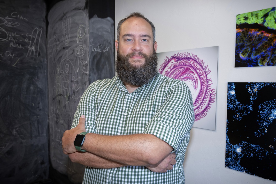 Portrait of a man in a checked shirt standing with his arms crossed in front of artwork. 