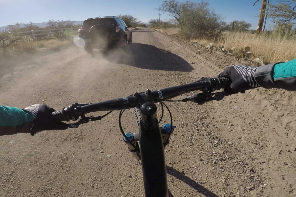 A vehicle kicks up dust as it passes a bicyclist on a dirt road