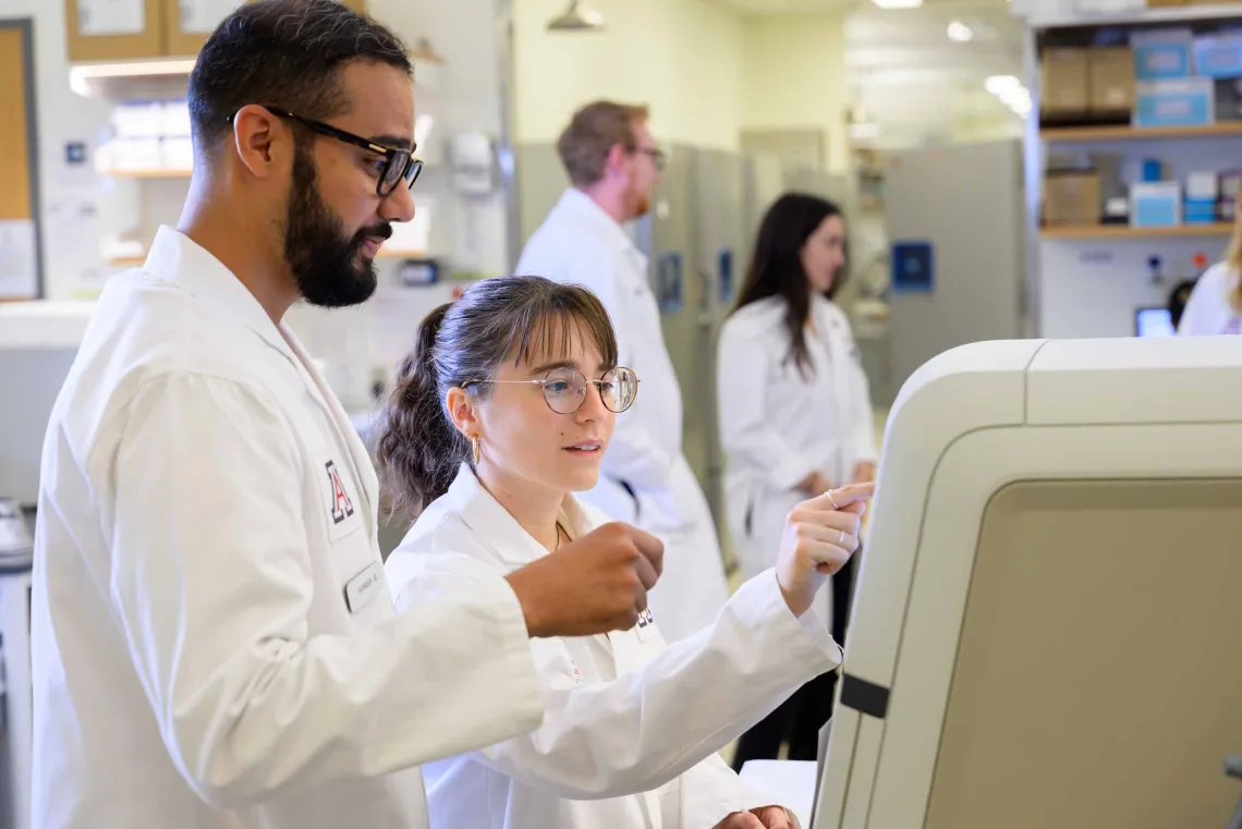 Two graduate students in white lab coats work on a large machine in a laboratory. 