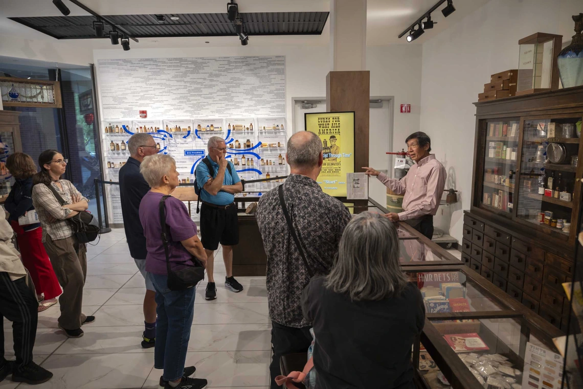 A group of about 10 people stands in the Coit Museum of Pharmacy and Health Sciences listening to Theodore Tong, PharmD, as he gives a tour. 