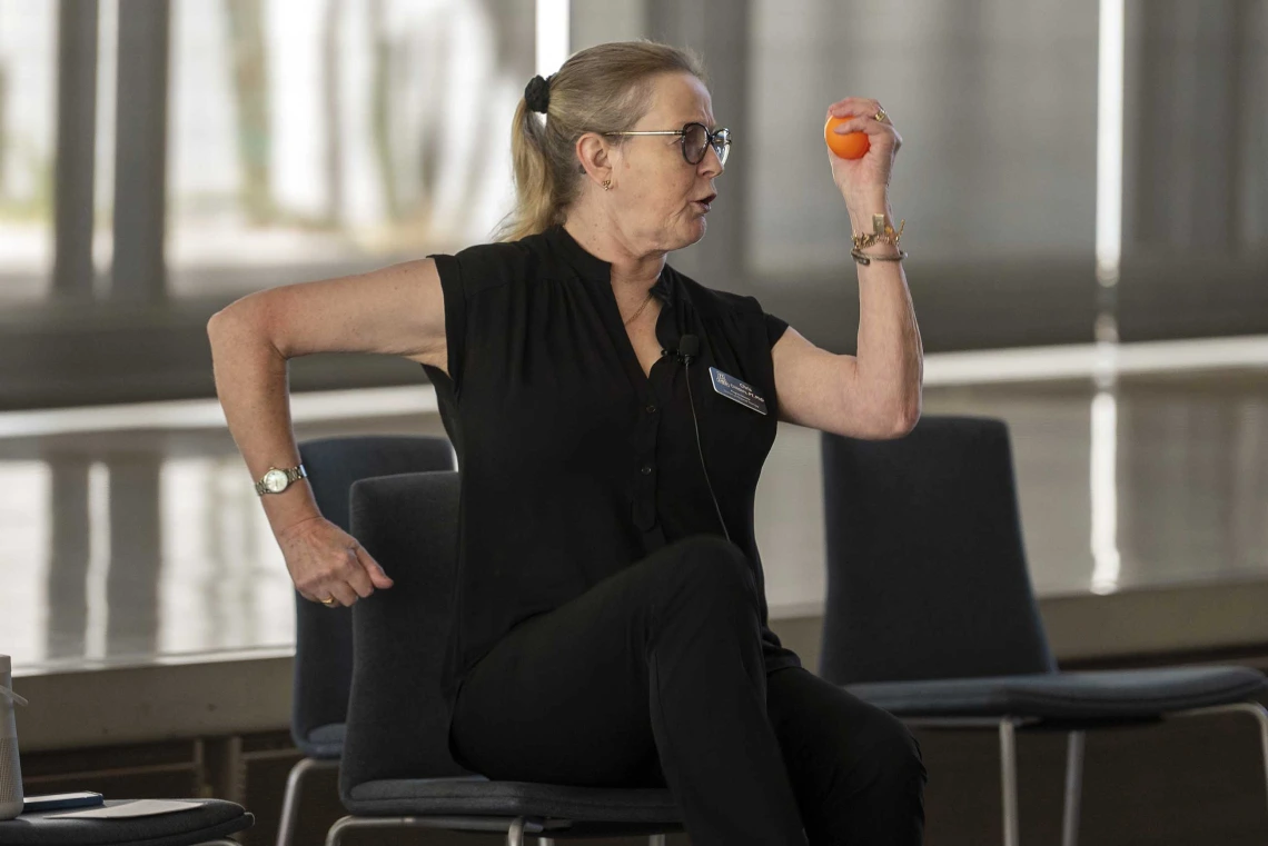 A woman sits in a chair with one arm back and the other forward, as if running in place. She holds a small ball in one hand. 