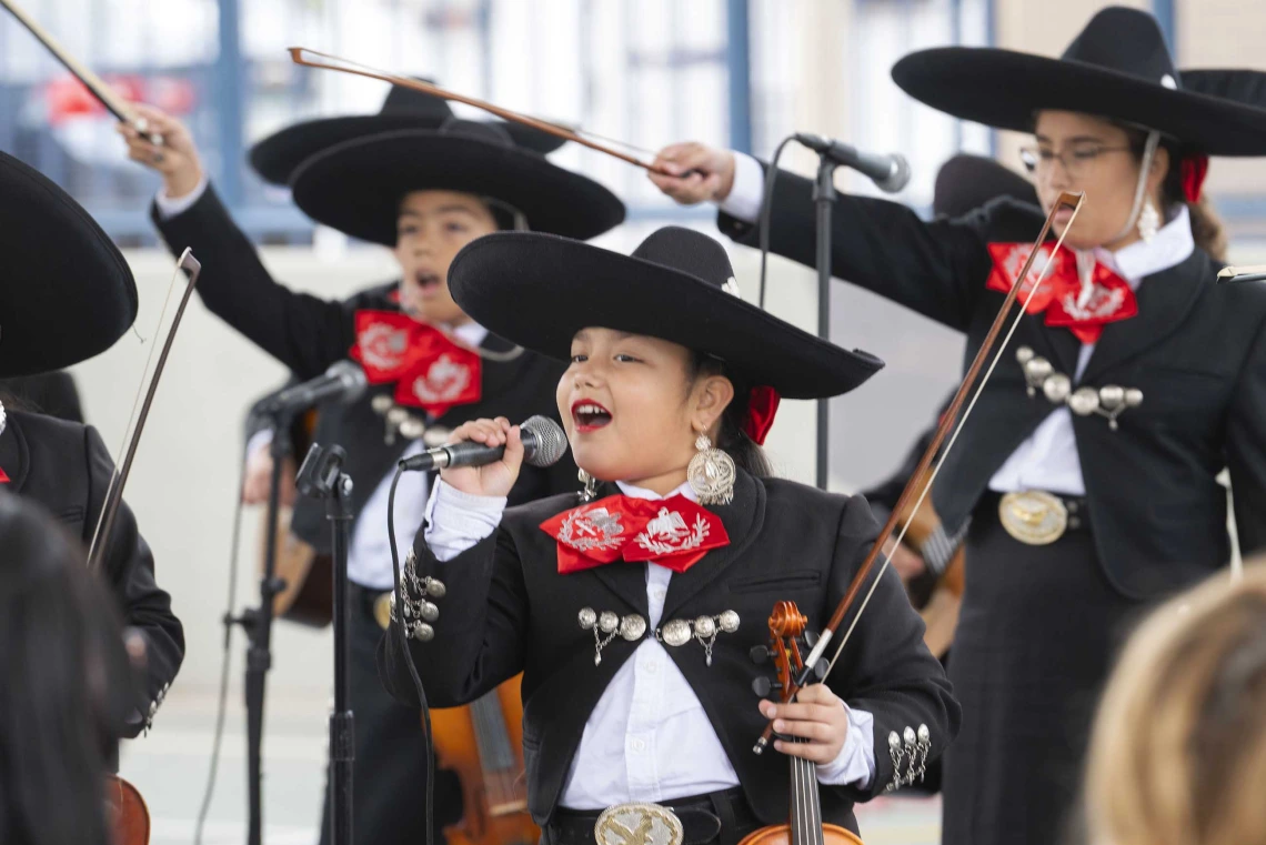 A young girl sings into a microphone in front of several other student mariachi musicians with violins who are all wearing black coats, pants and hats with red and white details. 