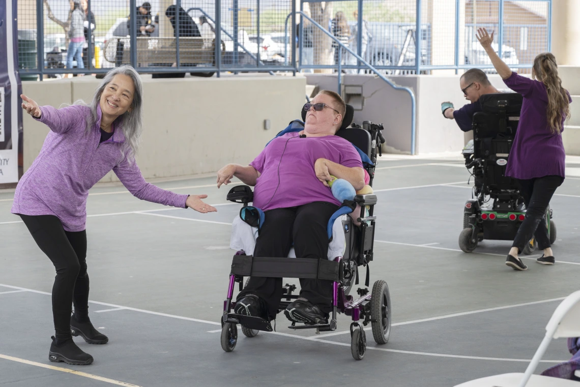 Two people using electric wheelchairs perform a dance with two people standing near them in an outdoor setting. 