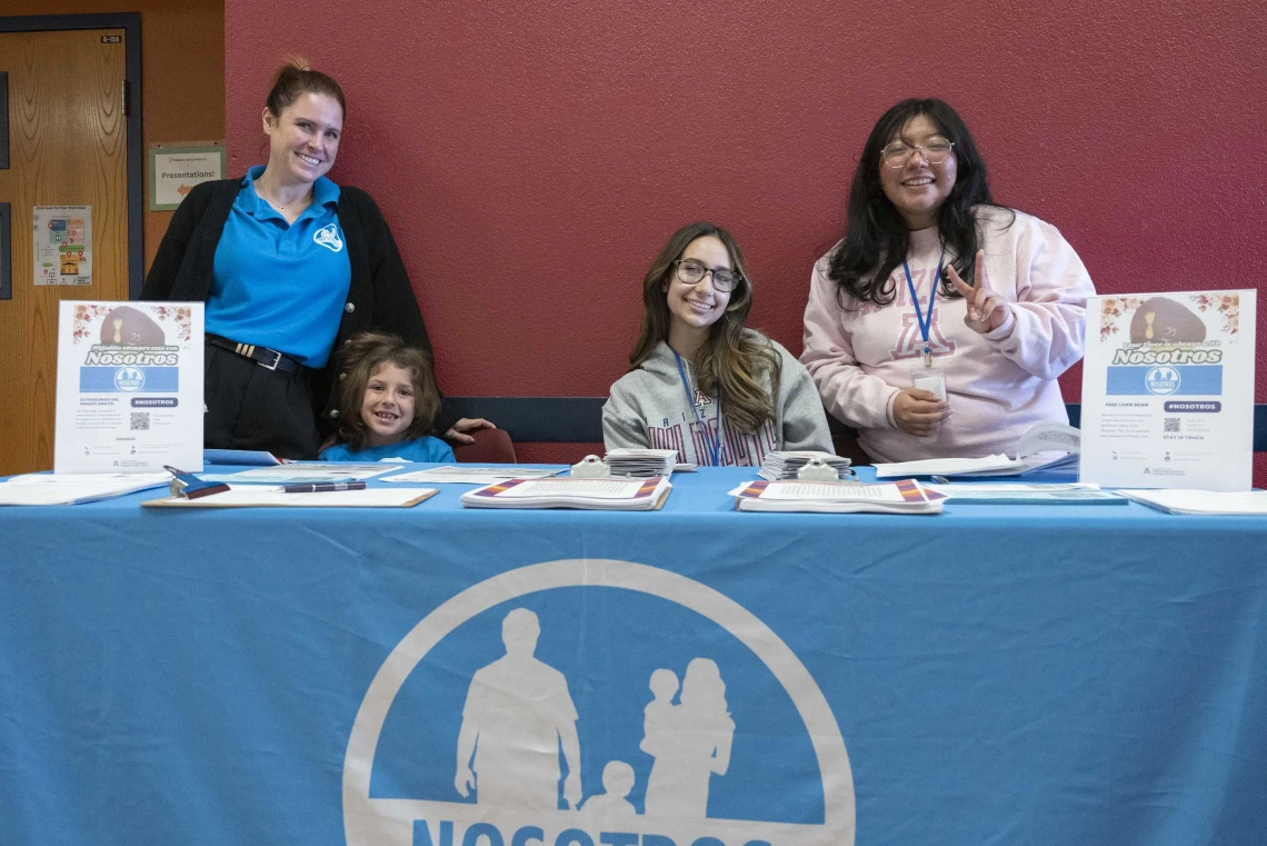 Three adults and a child stand behind a blue information table with some handouts and small posters. 