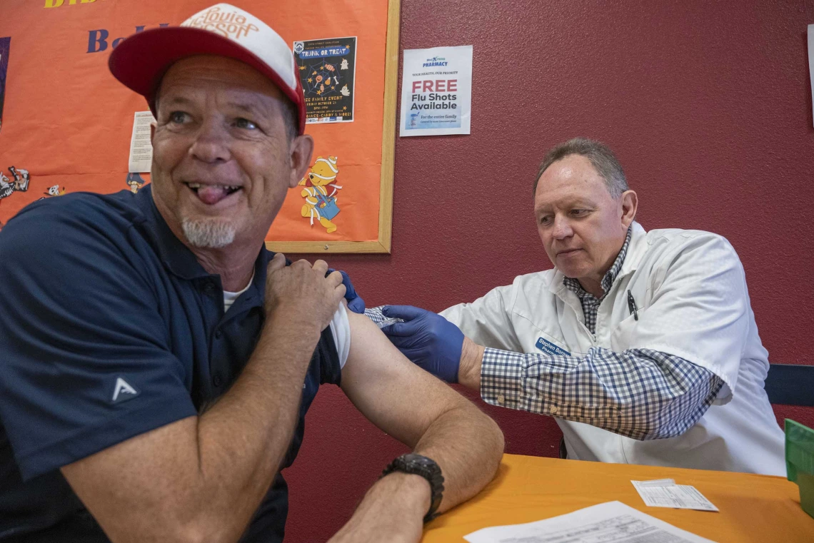 A man sitting at a table looks away and sticks out his tongue as a medical working gives him a shot in the arm.