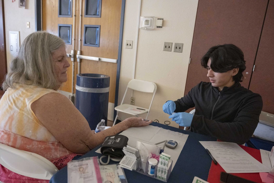 A woman sits at a table with her hand extended for a blood test being administered by a person wearing medical gloves. 