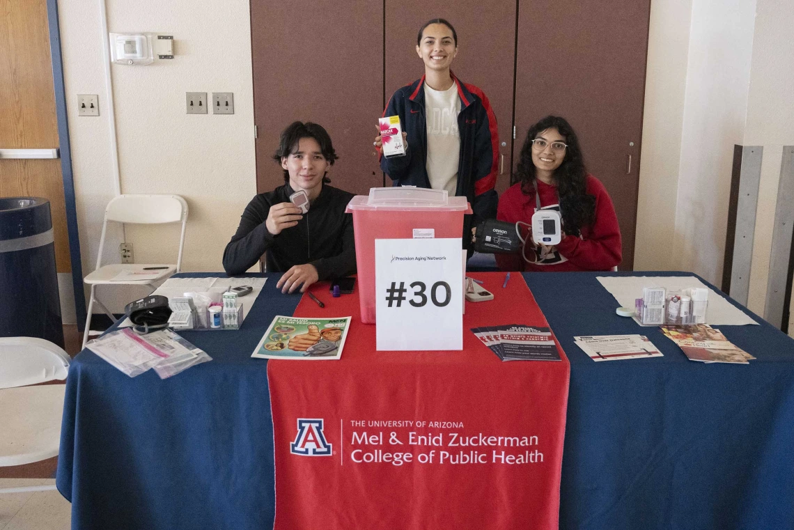 Three university students stand ready to give information at a table with a sign that reads “Mel and Enid Zuckerman College of Public Health.”