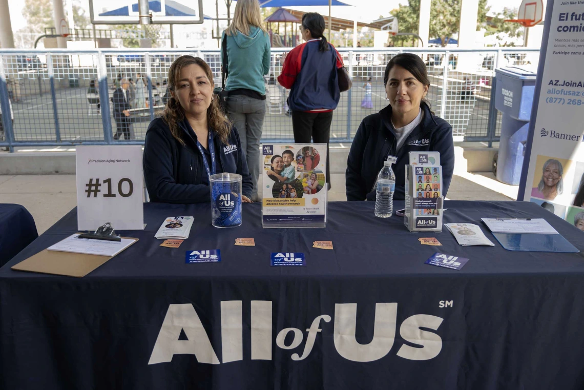 Two women stand behind an information table with a large sign that reads “All of Us.”