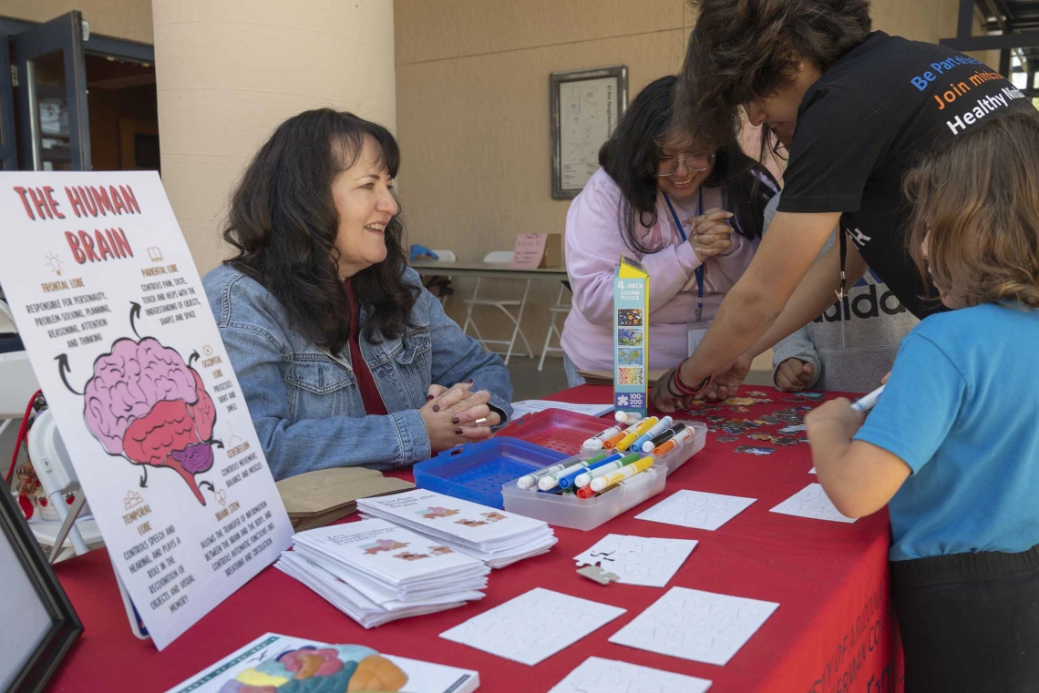 A woman sits at an information table covered with brochures and posters as people stand and talk with her. 