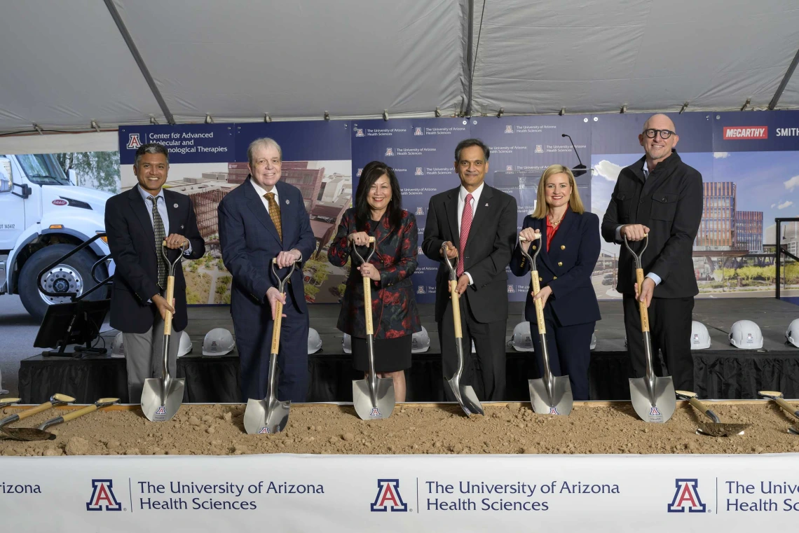 Six smiling people in suits and dresses hold shovels in sand with a University of Arizona Health Sciences banner in front of them.