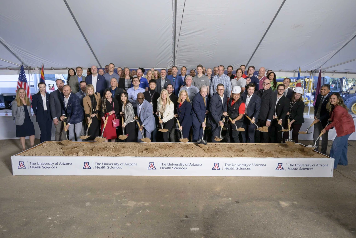 Several dozen people stand behind a large box of sand with shovels in hand. The banner across the front of the box reads “University of Arizona Health Sciences.”