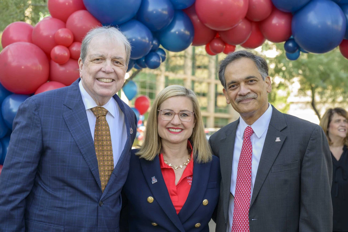 Two men in dark suits and ties stand on either side of a women in a dark blazer in an outdoor setting with red and blue balloons over their heads. All are smiling. 