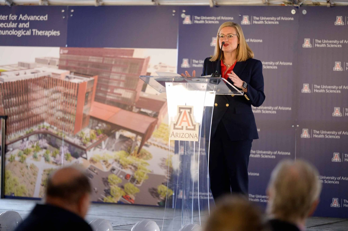 A women dressed in a dark pantsuit stands at a podium with the University of Arizona logo on it. There is a rendering of a building on a backdrop behind her. 